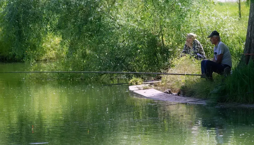 Dwaj wędkarze siedzą nad zieloną wodą, czekając na branie. Jeden z nich, starszy mężczyzna, zrelaksowany, z wędką w ręku, gotowy do **casting spinning**.